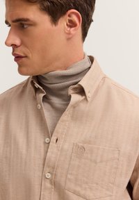 Mann in einem beigefarbenen Hemd mit Knöpfen über einem hellbraunen gerippten Rollkragenpullover, der nach links auf einem schlichten hellen Hintergrund blickt.