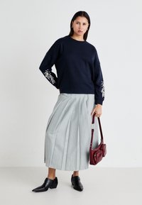 Sudadera navy con detalles florales bordados en las mangas, combinada con una falda plisada gris claro y botas de tobillo negras de tacón. Bolso burdeos.