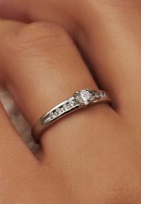 Bague en argent avec une pierre ronde centrale, accentuée par de plus petites pierres le long de l'anneau. Fini lisse et poli, posée sur une main à la peau claire.