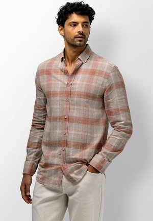 Mann trägt beige und rostfarben kariertes Hemd mit Knöpfen und hellbeige Hose, steht mit einer Hand in der Tasche und schaut zur Seite.