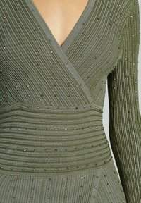 Rochie verde măsliniu cu decolteu în V, mâneci lungi, textură ribbed cu accente de mărgele și talie strânsă, care se transformă într-o fustă evazată.