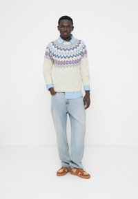 Πλεκτό πουλόβερ σε κρεμ απόχρωση με πολύχρωμα γεωμετρικά σχέδια, φορεμένο πάνω από ένα μπλε ριγέ πουκάμισο με γιακά, συνδυασμένο με ανοιχτό μπλε τζιν και καφέ παπούτσια.