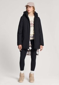 Kobieta w czarnej, zimowej kurtce z kapturem, beżowej czapce, kremowej bluzie z grafiką, koszuli w kratę związanej w talii, czarnych legginsach oraz beżowych zimowych butach.