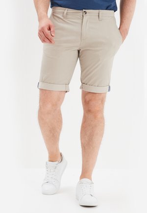 Man draagt beige opgerolde shorts, witte sneakers en een donkerblauw shirt, staat met één hand in zijn zak tegen een effen achtergrond.