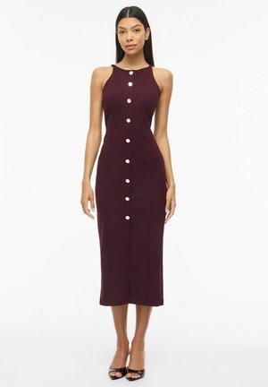 Kobieta stoi, ubrana w dopasowaną, bezrękawnikową sukienkę midi na guziki w ciemnym fioletowym kolorze oraz czarne sandały na obcasie z odkrytymi palcami na białym tle.