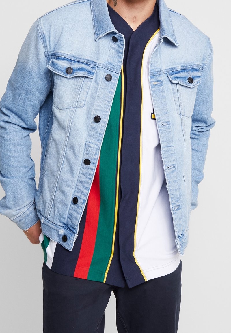 Ljusblå denimjacka med knappstängning framtill, två bröstfickor och en avslappnad passform. Under har man en flerfärgad randig t-shirt med marinblå bas.