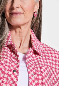 Roze en crèmekleurige houndstooth jasje met metalen drukknoopjes, gelaagd over een witte blouse, gecombineerd met bloemvormige hangende oorbellen.