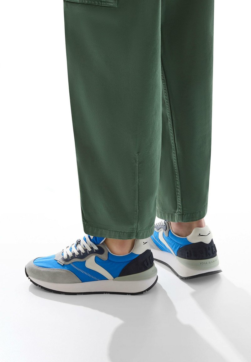 Sneakers blu e grigi con lacci bianchi e dettagli neri. Presentano una tomaia in morbido tessuto e una suola in gomma. Indossati con pantaloni verdi.