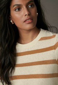 Cremefarbener Strickpullover mit horizontalen rostfarbenen Streifen, rundem Ausschnitt, gerippten Kanten und strukturiertem Stoff. Hat schulterlanges, dunkles Haar und trägt goldene Ohrringe.