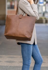 Sac cabas en cuir marron avec une texture lisse, une forme structurée et une poignée supérieure. Présente une ligne de couture discrète et un petit détail de logo.