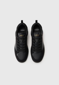 Ein Paar schwarze ARKK Copenhagen Sneakers mit Schnürsenkeln, sichtbarem Markennamen in Gold auf der Zunge, von oben auf einem weißen Hintergrund betrachtet.