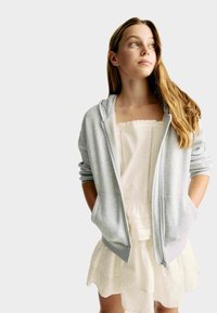 Grå zip-up hoodie med en mjuk textur och två framfickor, bärs över en vit lagad klänning med en volangkant och hålmönster.