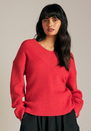 Maglione a coste rosso con scollo a V e maniche lunghe, caratterizzato da strisce testurizzate e una vestibilità morbida. Indossato sopra una gonna nera.
