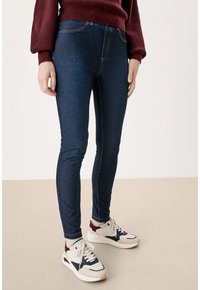 Tmavě modré denimové legíny s úzkým střihem, zdobené kontrastním prošíváním a vysokým pasem, v kombinaci s bílými a tmavě modrými teniskami.