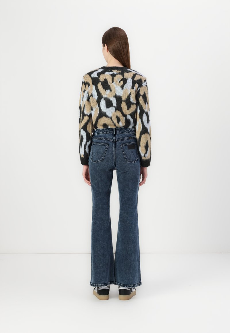 Strickpullover mit einem Leopardenmuster in Beige, Schwarz und Hellblau; kombiniert mit hochgeschnittenen, ausgestellten Jeans und Turnschuhen.