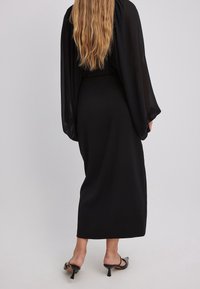 Vestido maxi negro con mangas largas transparentes y fluidas. Textura suave, cintura ajustada y diseño minimalista. Sandalias de tacón parcialmente visibles.