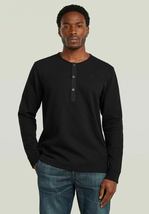 Mand iført sort langærmet henley-skjorte med tre knapper og blå jeans, stående med den ene hånd i lommen mod en ensfarvet baggrund.