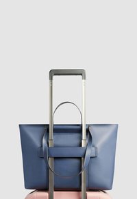 Blauwe leren shopper met twee handvatten, rustend op een roze koffer met een uitschuifbare zilveren handgreep. Gladde textuur en gestructureerd ontwerp.