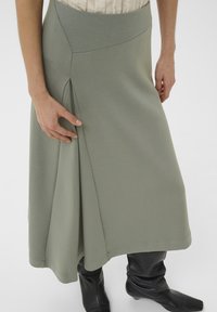 Hellgrüner Midirock mit glatter Textur, einem seitlichen Plissee-Detail und einem taillierten Bund, kombiniert mit schwarzen kniehohen Stiefeln.