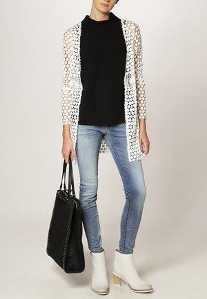 Cárdigan de ganchillo blanco sobre un cuello de tortuga negro, combinado con jeans de un azul claro y botines blancos de tacón. Incluye un bolso tote negro.