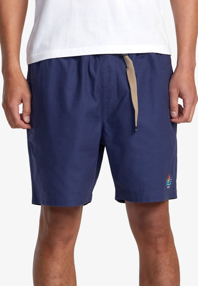 Meripuku navy sinikäy, jotka on valmistettu kevyestä kankaasta. Niissä on joustava vyötärö, beige nyöri ja pieni monivärinen logo helman lähellä.
