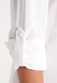 Manica di una camicia bianca con polsino abbottonato. Tessuto liscio e leggero; presenta un polsino piegato con un bordo arrotondato e un singolo bottone bianco.