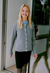 Grau Wollcardigan mit perlenartigen Knöpfen, getragen über einem blau-weiß gestreiften Hemd. Kombiniert mit einem schwarzen Rock, steht sie neben einer Tür.