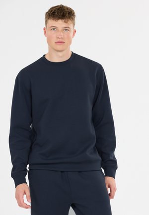 Ung mand med kort krøllet hår iført en ensfarvet mørk navy sweatshirt og matchende bukser står mod en hvid baggrund.