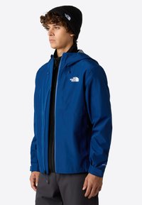 Modrá voděodolná bunda s kapucí, která má přední zip, elastické manžety a logo na hrudi. Nositá přes černý top.