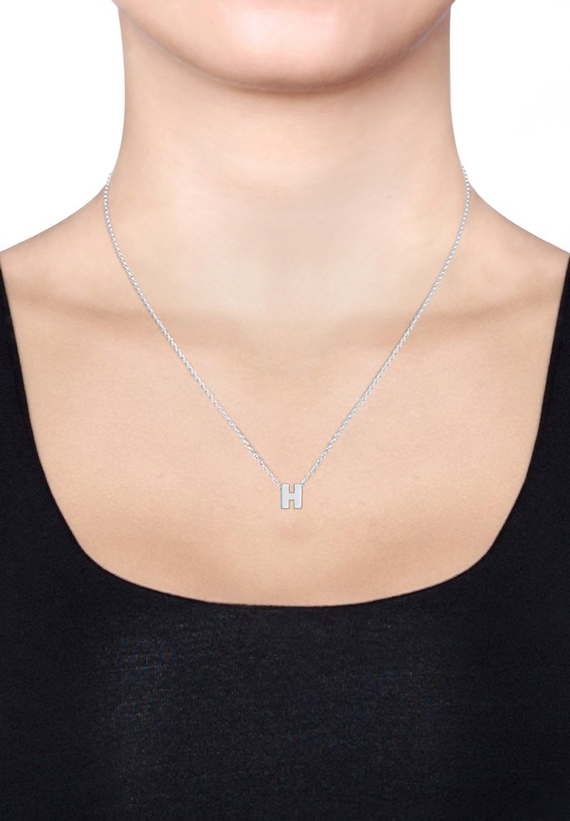 Collier en argent avec un simple pendentif en forme de « H » sur une chaîne délicate, présenté sur un haut noir et un fond neutre.