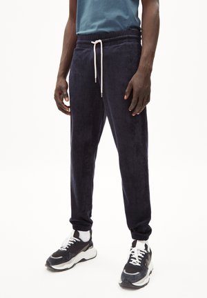 Man draagt donkerblauwe joggers met elastische boorden, een witte aantrekkoord, witte sokken en zwart-witte sneakers tegen een effen achtergrond.