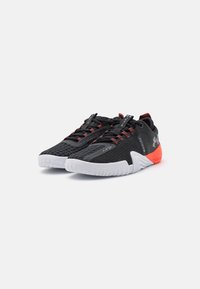 Sorte sportssko med røde detaljer, mesh-overdel, struktureret design, hvid gummisål og rød hældetalje; snørebånds lukning.