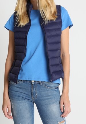 Kvinde iført en blå kortærmet skjorte, navy dunvest og lyseblå revnede jeans, stående mod en ensfarvet baggrund.