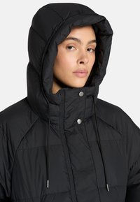 Zwarte pufferjas met een grote capuchon, voorzien van een gestikt patroon, trekkoorden en drukknoopsluiting. Zacht, lichtgewicht materiaal.