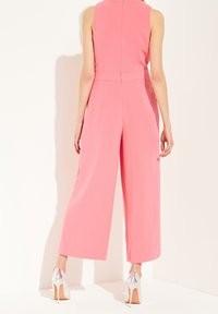 Růžový overal bez rukávů se širokými nohavicemi, strukturovanou látkou a zapínáním na zip na zádech. Kombinován se stříbrnými lodičkami na vysokém podpatku.