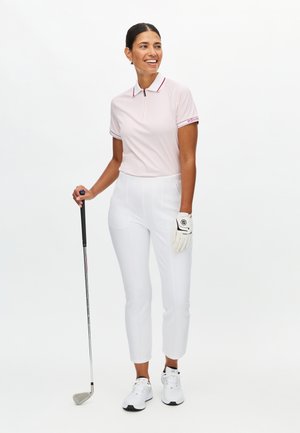 Jasnoróżowa koszulka polo do golfa z krótkim rękawem i czerwonymi akcentami, zestawiona z białymi spodniami golfowymi. Model trzyma kij golfowy i ma na sobie białą rękawiczkę oraz trampki.