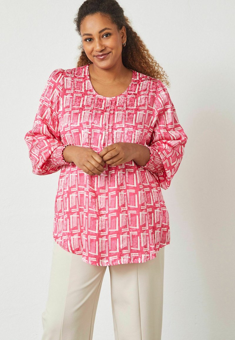 Femme aux cheveux bouclés portant un chemisier à manches longues rose et blanc à motifs et un pantalon crème, debout devant un fond clair uni.
