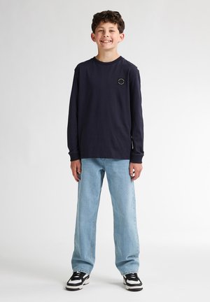 Marineblaues Langarmshirt mit Rundhalsausschnitt und kleinem Logo, kombiniert mit hellblauen, lässig geschnittenen Jeans und schwarz-weißen Sneakers.
