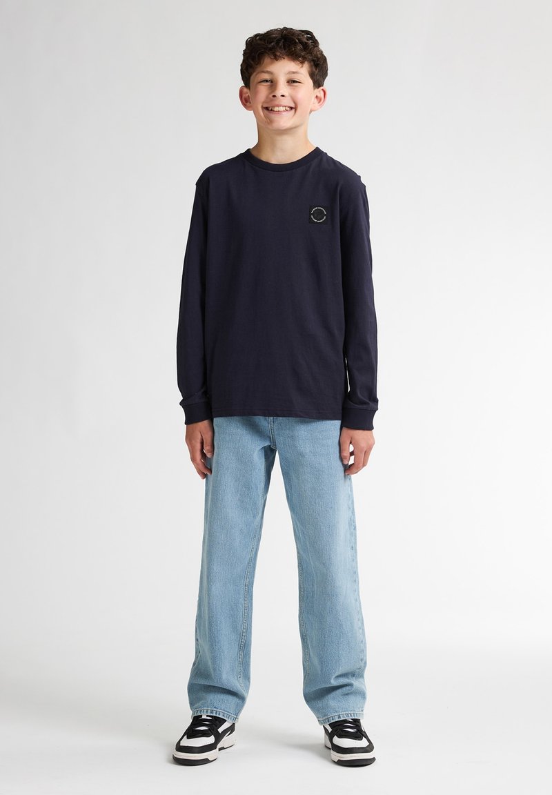 Camisa de manga larga azul marino con cuello redondo y un pequeño logo, combinada con jeans de corte holgado en azul claro y zapatillas negras y blancas.