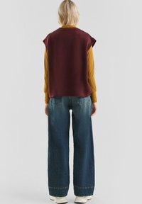 Vrouw met kort blond haar, draagt een bordeauxrode mouwloze trui over een mosterdgele ribbelige lange mouwtop, wijdvallende blauwe jeans en witte schoenen.