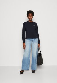 Maglietta a maniche lunghe navy con logo bianco, abbinata a jeans a vita alta di un azzurro chiaro con gamba larga e scarpe flat nere, mentre tiene una borsa a tracolla nera intrecciata.