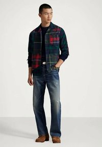 Man wearing dark plaid jacket, navy shirt, blue jeans, and brown shoes, standing with one hand in pocket, looking sideways.