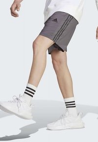 Άτομο που φοράει γκρι σορτσάκι Adidas, λευκές αθλητικές κάλτσες με μαύρες ρίγες και λευκά αθλητικά παπούτσια, περπατάει με το ένα πόδι υψωμένο.