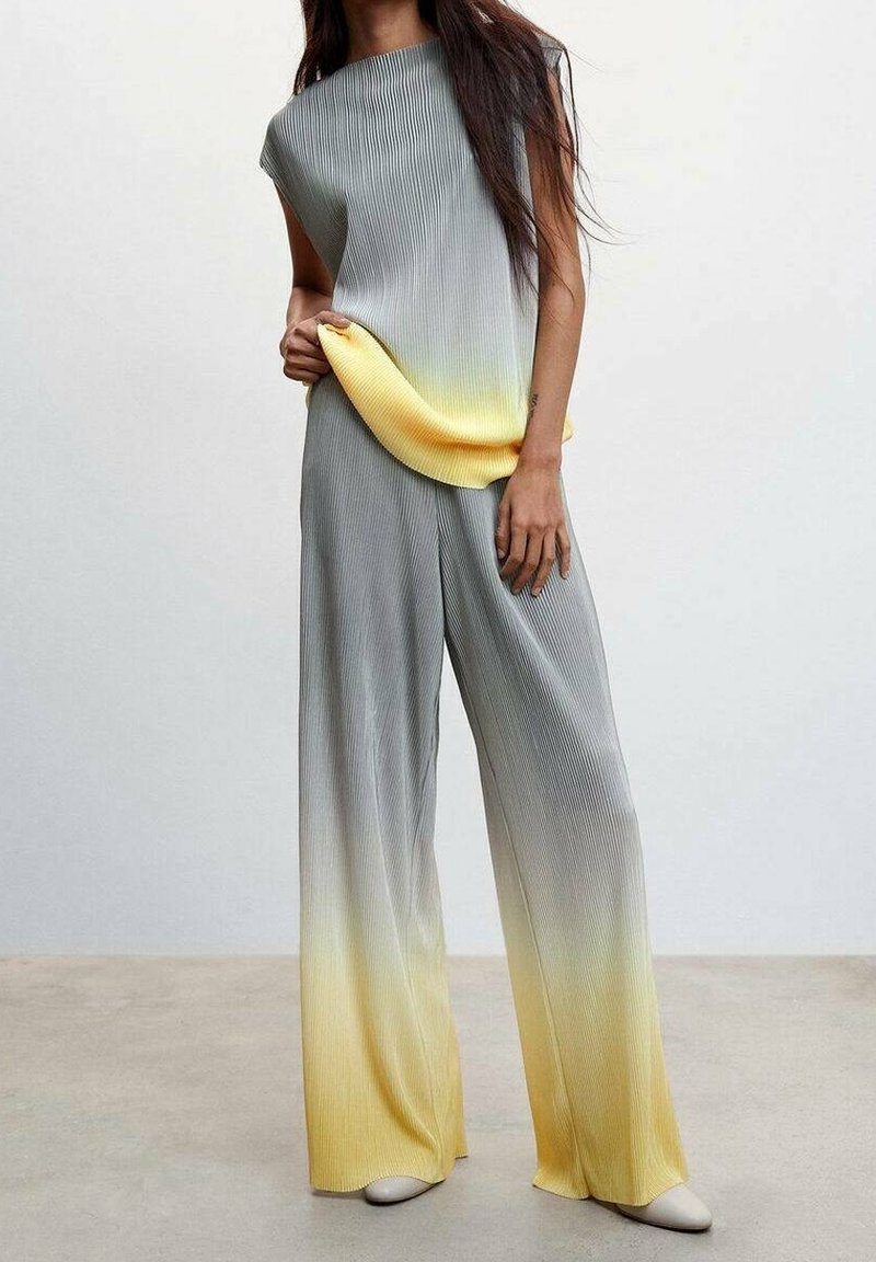 Femme portant un top sans manches côtelé gris et jaune en dégradé et un pantalon à jambes larges, debout sur un sol lisse devant un fond blanc uni.