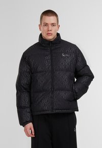 Giacca a vento nera con motivo trapuntato, colletto alto e chiusura con zip. Il tessuto ha una leggera lucentezza e un logo in evidenza sul petto.