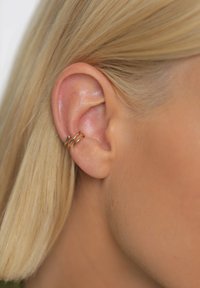 Náušnica v tvare kruhu v zlatej farbe so tromi prepletenými krúžkami. Detailný pohľad zdôrazňuje hladkú kovovú textúru na svetlej pokožke.