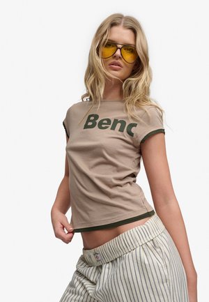 Ung kvinde med blondt hår iført gule tonede briller, en beige T-shirt med teksten "Benc" og stribede løse bukser mod en hvid baggrund.