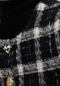 Sort-hvidternet stof med en tekstureret overflade; har en guldknap og en kontrasterende sort turtleneck under.