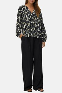 Langärmlige Bluse mit einem abstrakten Muster aus schwarzen, weißen und olivgrünen Blättern. V-Ausschnitt-Design, lockere Passform, kombiniert mit weiten schwarzen Hosen.