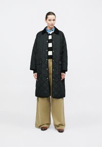 Czarny pikowany długi płaszcz z kołnierzem, noszony na paskowanym swetrze i szerokich, beżowych spodniach, z zatrzaskami i bocznymi kieszeniami.